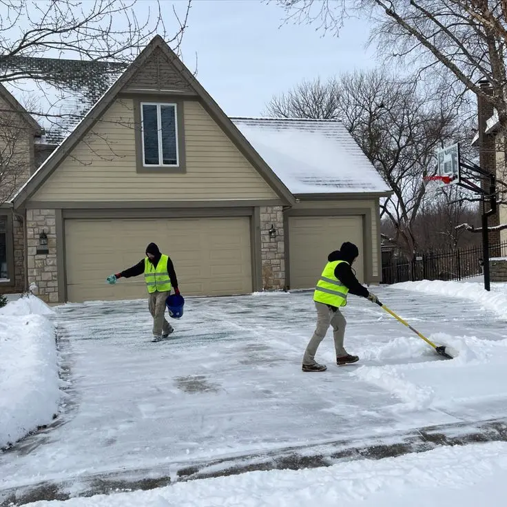 Two crew members applying rock salt and clearing ice from a home driveway in Brown County to prevent slip injuries.
