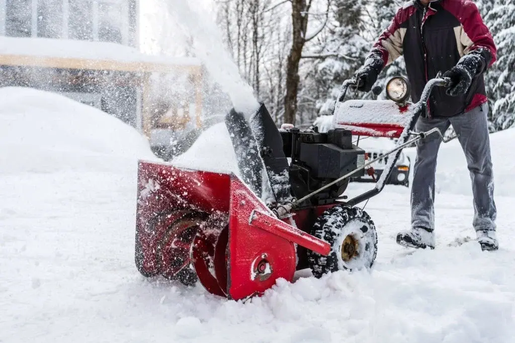 Close-up of a worker operating a gas-powered red snow blower throwing fresh snow off a residential property pathway.